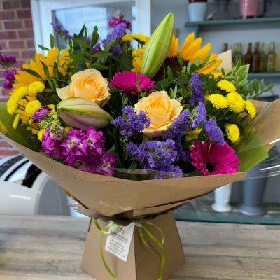Sunshine Surprise - A bold and joyful hand-tied bouquet featuring sunflowers, peach roses, lilac stock, gerbera, and bright yellow button chrysanthemums. A guaranteed day-brightener in a kraft box.